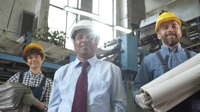 Zoom in of Caucasian male and female workers in overalls and hardhats holding newspapers and paper reels and Latin American manager standing together in printing plant and smiling at camera