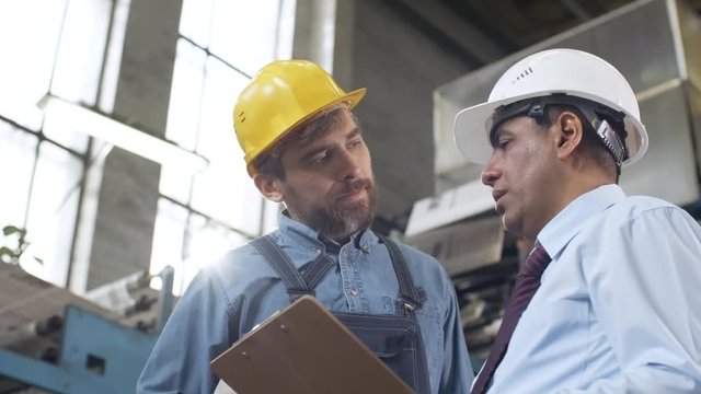 Low angled panning shot of middle aged Latin American manager in hardhat explaining something on clipboard and giving instructions to Caucasian male technician in factory