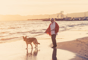 Happy attractive senior woman with her german shepard dog walking on the beach at autumn sunset