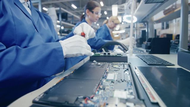 Close Up of a Female Electronics Factory Worker in Blue Work Coat Assembling Laptop's Motherboard with a Screwdriver. High Tech Factory Facility with Multiple Employees. 