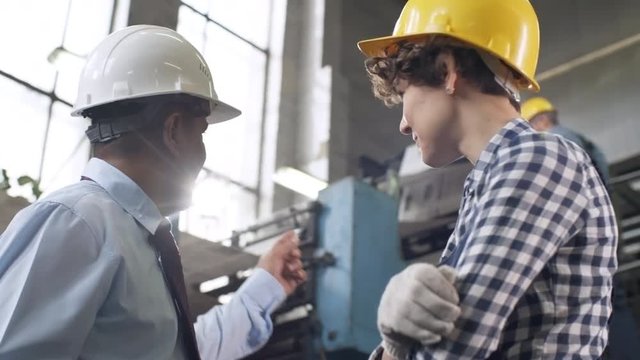Low angled panning shot of middle aged Latin American manager in hardhat and young female technician smiling and discussing document on clipboard while working together in industrial factory