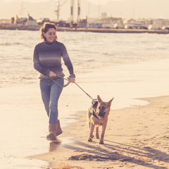 Happy attractive young woman with her german shepard dog walking on the beach at autumn sunset