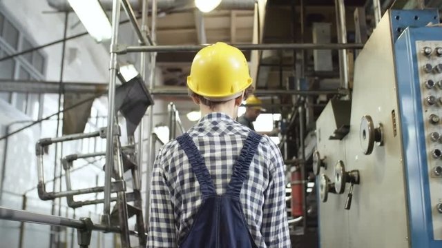 Following shot of female worker in overall and hardhat walking through industrial plant and looking around