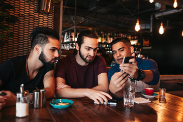 Group of mixed race young men talking and using phone in lounge bar. Multiracial friends having fun in cafe