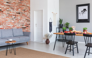 Grey sofa against red brick wall in flat interior with poster and black chairs at dining table. Real photo