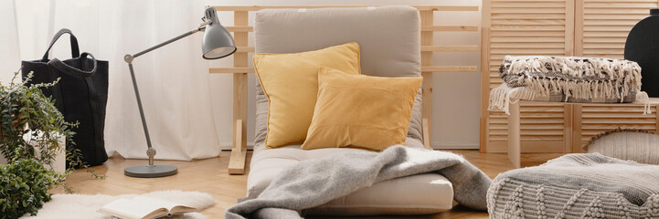 Two yellow pillows placed on futon mattress chair in real photo of bright living room interior with metal lamp, fresh plants, natural materials and book on the floor