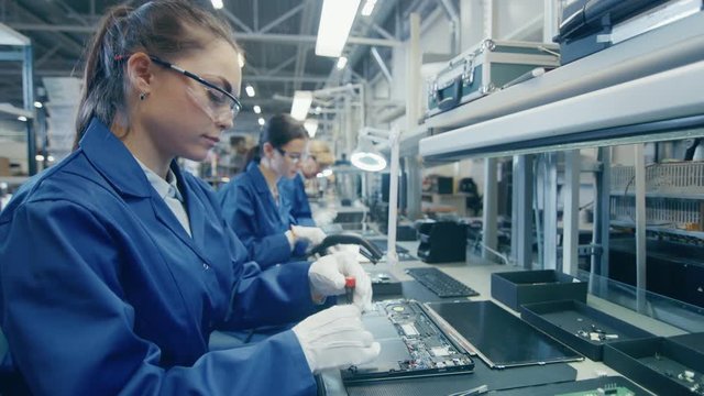 Woman Electronics Factory Worker in Blue Work Coat and Protective Glasses is Assembling Laptop's Motherboard with a Screwdriver. High Tech Factory Facility with Multiple Employees. 
