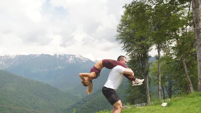 Athletic woman doing single leg split squat exercise with her male partner outdoors over high mountains in background