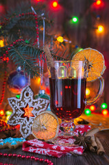 A glass of mulled wine with cinnamon and a slice of orange is on the table against the background of Christmas tree decorations on the branches of the Christmas tree in the light of Christmas lights i