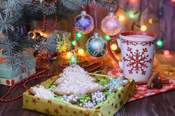 Obraz na płótnie Canvas Gingerbreads in the form of Christmas tree and stars in a gift box are under the Christmas tree on the table next to a glass of cocoa in the light of Christmas lights in the background