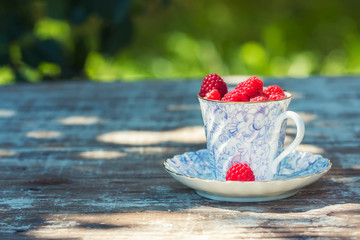 Fresh ripe raspberries and an old porcelain cup with a saucer on a wooden table in the garden.