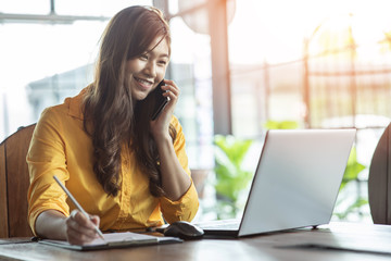 Beautiful Attractive Asian woman working with laptop smile and talking with customer in smartphone to get idea and requirement for success job outside office in coffee shop,Business Concept,Warm Tone