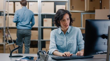 Female Inventory Manager Sitting at Her Desk and Using Personal Computer, Worker Puts Package on the Designated Shelf. In the Background Rows of Cardboard Boxes with Products Ready For Shipment.