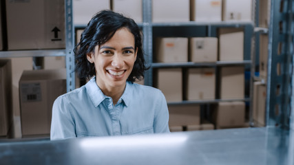 Female Warehouse Worker Smiles Charmingly at the Camera. In the Background Rows of Shelves Full of...
