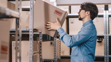 Warehouse Worker Puts Cardboard Box on a Shelf. In the Background Rows of Shelves Full of Cardboard...
