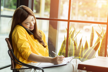 Beautiful Attractive Asian Business woman writing down requirement to book and talking with customer in smartphone,Feeling so happiness and working with service mind,Business and Finance Concept