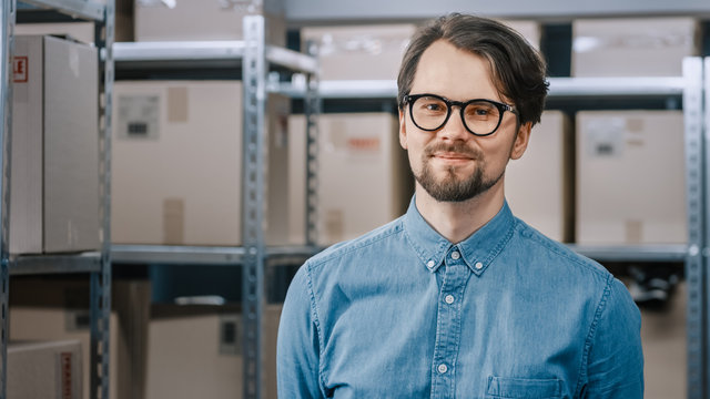 Portrait Of The Handsome Warehouse Inventory Manager Standing And Smiling At The Camera. Smart Man Wearing Glasses With Rows Of Shelves Full Of Cardboard Boxes And Parcels Ready For Shipment.