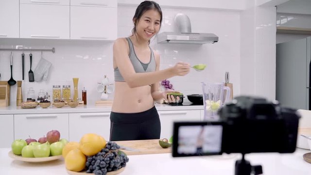 Blogger sporty Asian woman using camera recording how to make avocado juice video for her subscriber, female use organic fruit making avocado juice by herself at home. Healthy food concept.