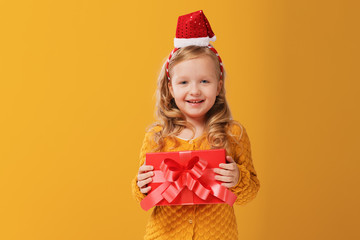 Happy little child girl in a warm knitted sweater and cap Santa Claus is holding a red box with a gift on a yellow  isolated background. The concept of celebration, giving and receiving a gift