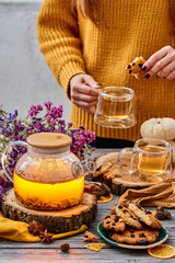 Girl in a yellow sweater holding a transparent cup of tea. Transparent teapot, cookies with cranberries and two transparent cups of tea on wooden cuts and dried flowers on the background. close up