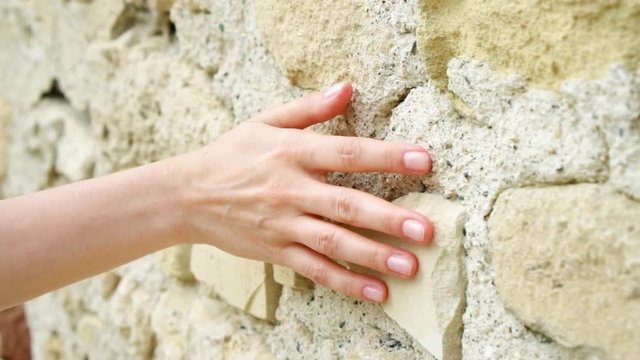 Woman sliding hand against old ancient stone wall in slow motion. Female hand touching hard rough surface of rock on sunny summer day