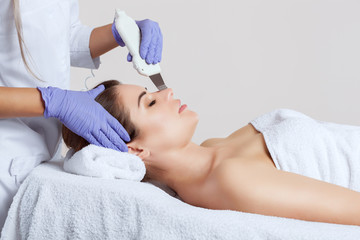 The doctor-cosmetologist makes the ultrasound cleaning procedure of the facial skin of a beautiful,...