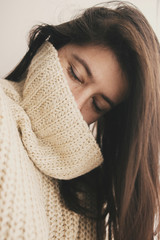 Stylish hipster girl in knitted sweater smiling in soft light in room. Portrait of young attractive woman relaxing and taking selfie. Atmospheric cozy winter moments. Moody image