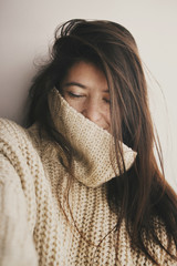 Portrait of young attractive woman with no makeup in knitted sweater smiling in soft light in room. Stylish hipster girl taking selfie. Atmospheric cozy winter moments. Moody image, carefree