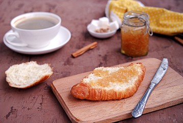 Breakfast: a cup of coffee, fresh croissant on a wooden board, smeared with orange jam.
