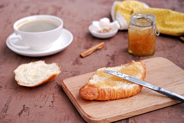 Breakfast: a cup of coffee, fresh croissant on a wooden board, smeared with orange jam.