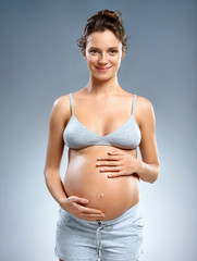 Happy pregnant woman touching her belly on grey background. Pregnancy, maternity, preparation and expectation concept