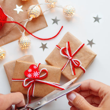 Woman is packing Christmas and New Year DIY presents in craft paper. Gifts tied with white and red threads. Boxes and star confetti on white background.