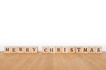 Focus on the phrase "Merry Christmas" made of wooden block dice with letters on a wooden table. Shallow depth of field. Copy space.