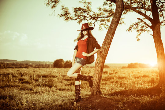 pretty cowgirl in hat and shirts leaning to tree at rural sunset landscape
