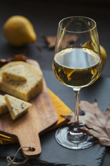 A glass of white wine served with cheese in a cutting board on dark background