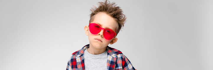 A handsome boy in a plaid shirt, gray shirt and jeans stands on a gray background. Boy in red sunglasses