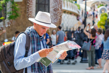 A young man in a scarf and hat with a backpack is holding a map in front at the entrance in Guell Park, Barcelona, Spain.