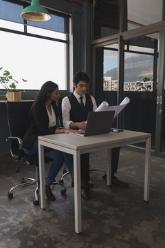 Executives discussing over blue print while using laptop