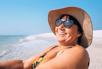 Happy smiling Elderly womanr woman in sunglasses and big hat takes sun bath on seaside