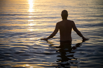 Sunrise silhouette of man standing in calm ocean waters facing the golden morning sun