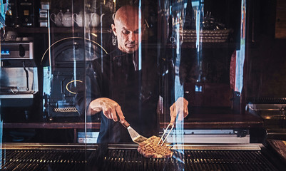 Chef cooking delicious beef steak on a kitchen, standing behind protective glass in a restaurant.