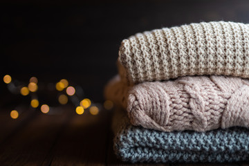 Obraz na płótnie Canvas Stack of cozy knitted sweaters on dark wooden background with light bokeh. Autumn-winter concept,knitted wool sweaters. Pile of knitted winter clothes, sweaters, knitwear, space for text.
