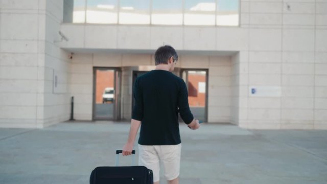 Young man pulling suitcase in modern airport terminal. Travelling guy wearing smart casual style clothes walking away with his luggage while waiting for transport. Rear view.