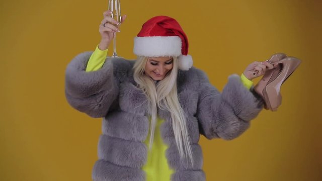 Christmas gifts, surprises or purchases. A young blond woman in a fur coat and Santa Claus hat is holding a pair of shoes and a glass of champagne. Happily dances from winning the Christmas lottery