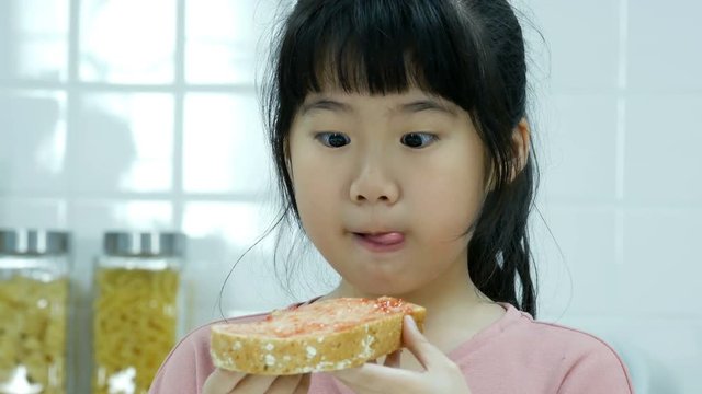 Close-up face little girl while eating bread with happy emotion. Asian girl eating bread at kitchen.