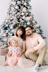 happy young family with cute baby girl holding sitting in front of the christmas tree and smiling at camera