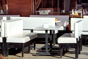 A wooden white table with white sofas on black legs against the background of the facade with windows of an outdoor cafe with empty seats on a sunny day.