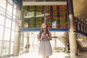 Theme travel and tranosport. Beautiful young caucasian woman in dress and backpack standing inside train station or terminal looking at a schedule holding a red phone, uses communication technology