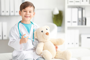Cute little boy dressed as doctor playing with toy bear in hospital