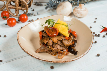 fried meat closeup on brown plate on white textured table with cherry tomatoes and chilli and pepper spices and green at background